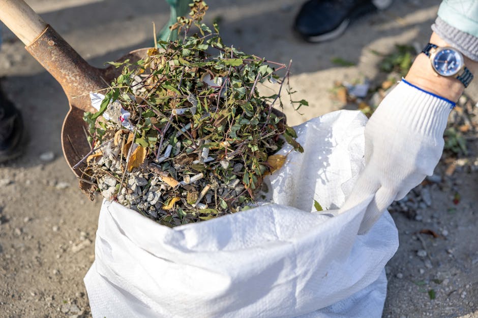 A white plastic bucket filled with small branches, twigs, and wood debris, placed on a grassy area adjacent to a large pile of chopped firewood and branches in a garden or yard setting. The bucket sho
