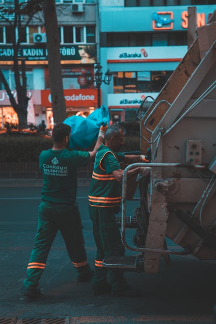 Two waste collection workers wearing dark green uniforms with reflective orange stripes are engaged in rubbish removal on a city street. One worker, standing to the left, is lifting a large blue bag f