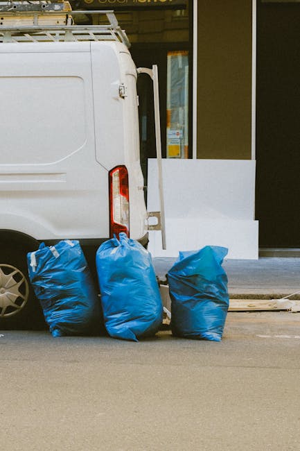 Two waste collection workers wearing dark green uniforms with reflective orange stripes are engaged in rubbish removal on a city street. One worker, standing to the left, is lifting a large blue bag f