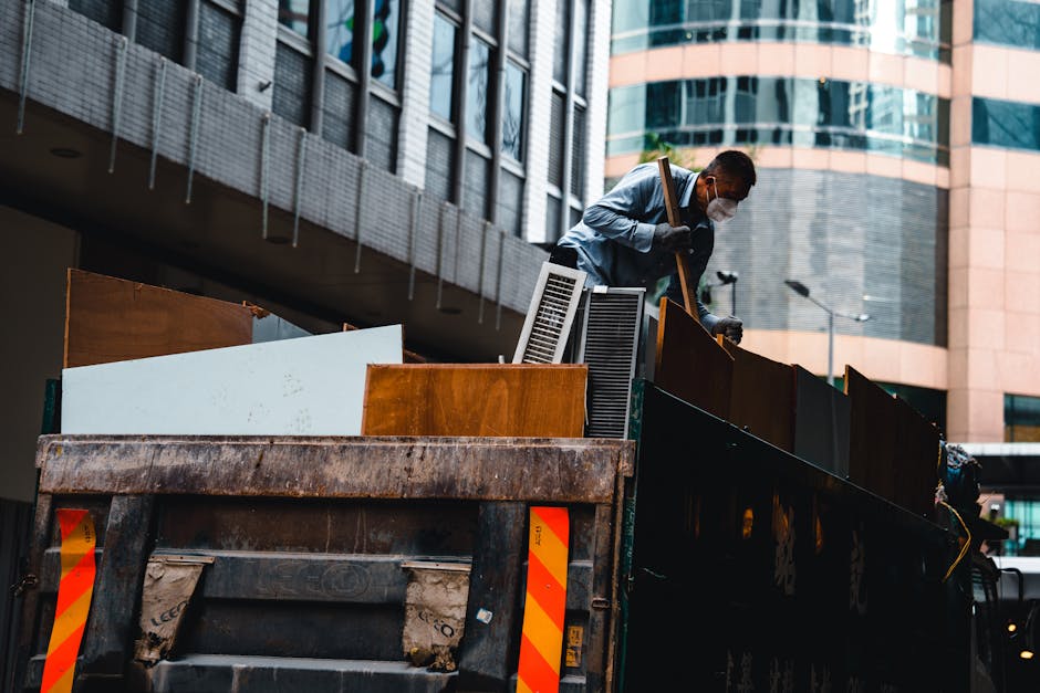 A worker wearing a face mask and gloves is standing on the edge of a large, dark green industrial waste container mounted on a flatbed truck, surrounded by a variety of bulky household items and furni